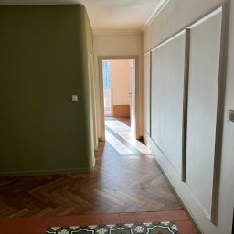 Jarosław Nitychoruk - Korytarz po remoncie z parkietem ułożonym w jodełkę, oliwkową ścianą i białą ścianą z panelami dekoracyjnymi, widok na dalsze pomieszczenia.