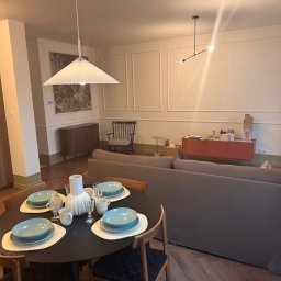 Jarosław Nitychoruk - Salon z parkietem w jodełkę, nakrytym stołem i szarą sofą na tle ścian z dekoracyjnymi listwami i pomalowanymi na kremowo. Częściowo widoczna szafka kuchenna po lewej stronie.