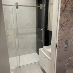 Nowoczesna łazienka z kabiną prysznicową ze szklanymi drzwiami przesuwnymi, marmurowymi płytkami, białą szafką umywalkową i tapetą z motywem drzewa.