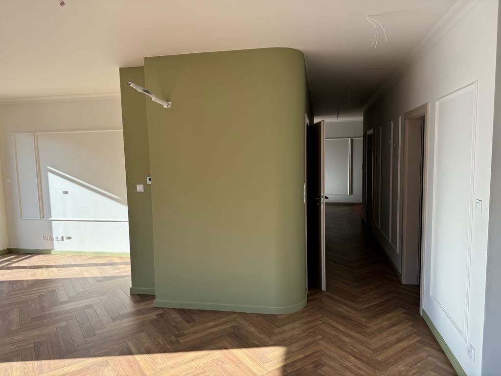 Wnętrze po remoncie z parkietem ułożonym w jodełkę, ścianą w odcieniu oliwkowej zieleni i białymi listwami przypodłogowymi. Widoczne niedokończone podłączenie przewodów elektrycznych.