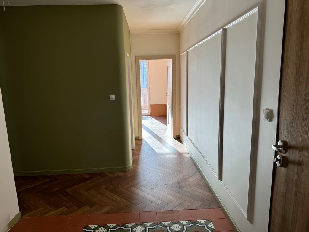Korytarz po remoncie z parkietem ułożonym w jodełkę, oliwkową ścianą i białą ścianą z panelami dekoracyjnymi, widok na dalsze pomieszczenia.