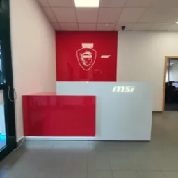 Nowoczesna recepcja w biurze z białym blatem i czerwonym panelem bocznym, logo firmy MSI na ścianie i blacie, widok na salę z pracownikami w tle.