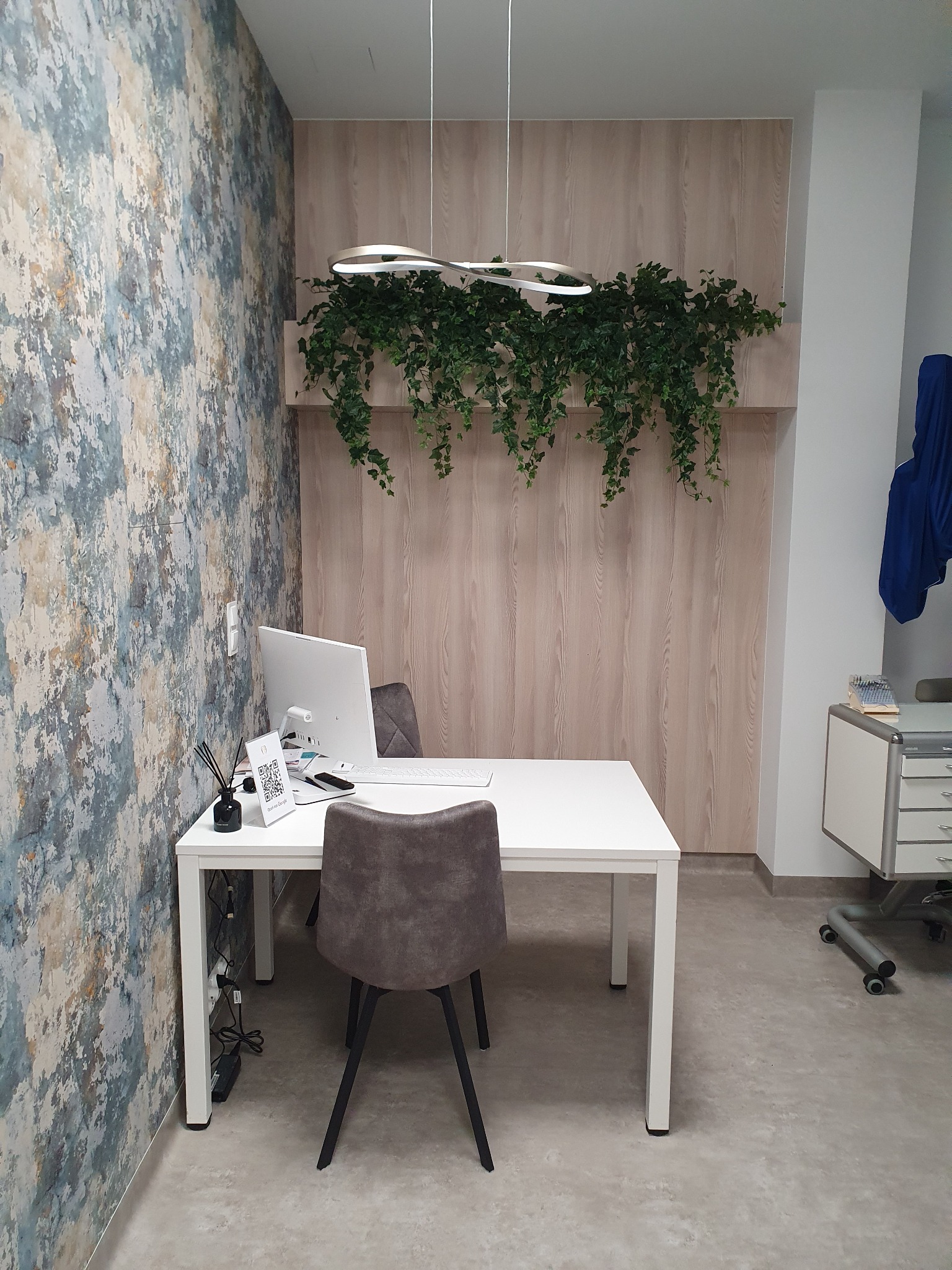 Nowoczesne biuro z białym biurkiem, szarymi krzesłami, dekoracyjną lampą i zielonymi roślinami zwisającymi ze ściany, obok biurka stoi biała szafka na kółkach.