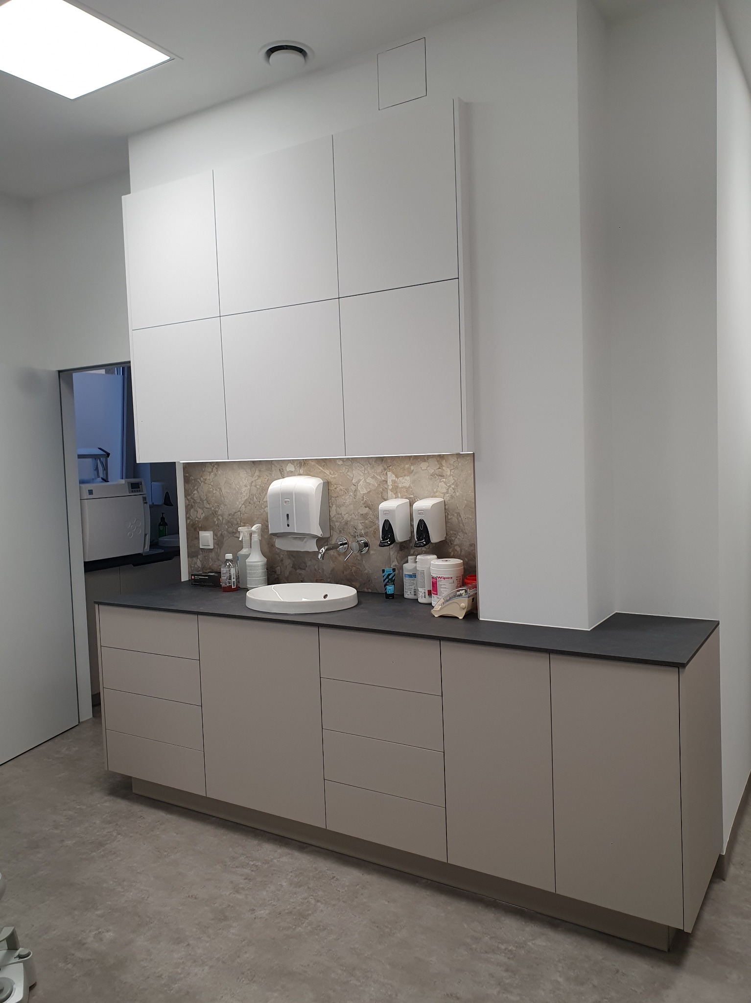 Nowoczesna zabudowa meblowa w gabinecie lekarskim z umywalką, dozownikami mydła i szafkami wiszącymi, oświetlona listwą LED.