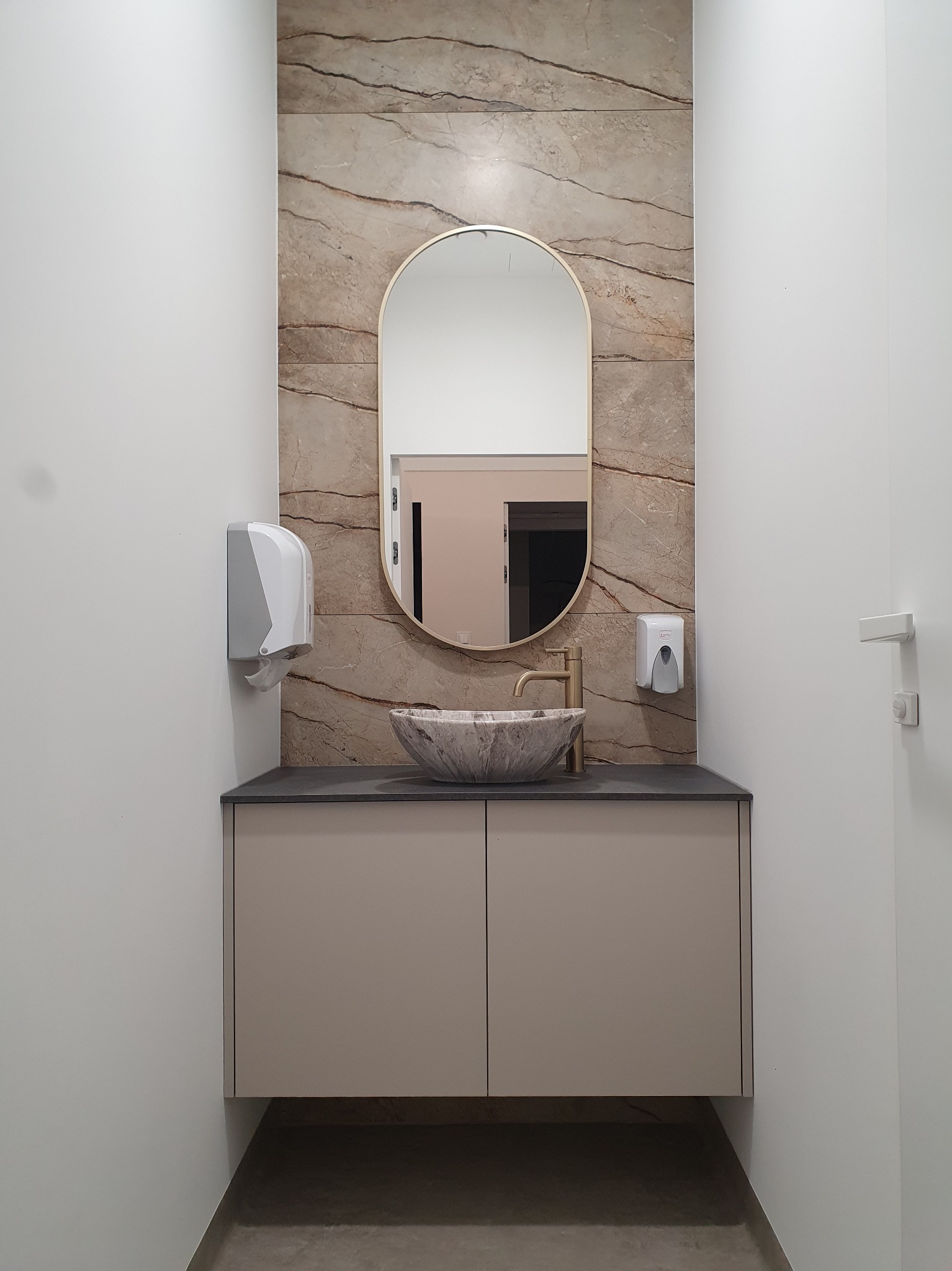 Nowoczesna łazienka z umywalką w kształcie misy z marmurowym wzorem, złotą armaturą i owalnym lustrem w złotej ramie, na tle ściany wyłożonej płytkami imitującymi marmur.