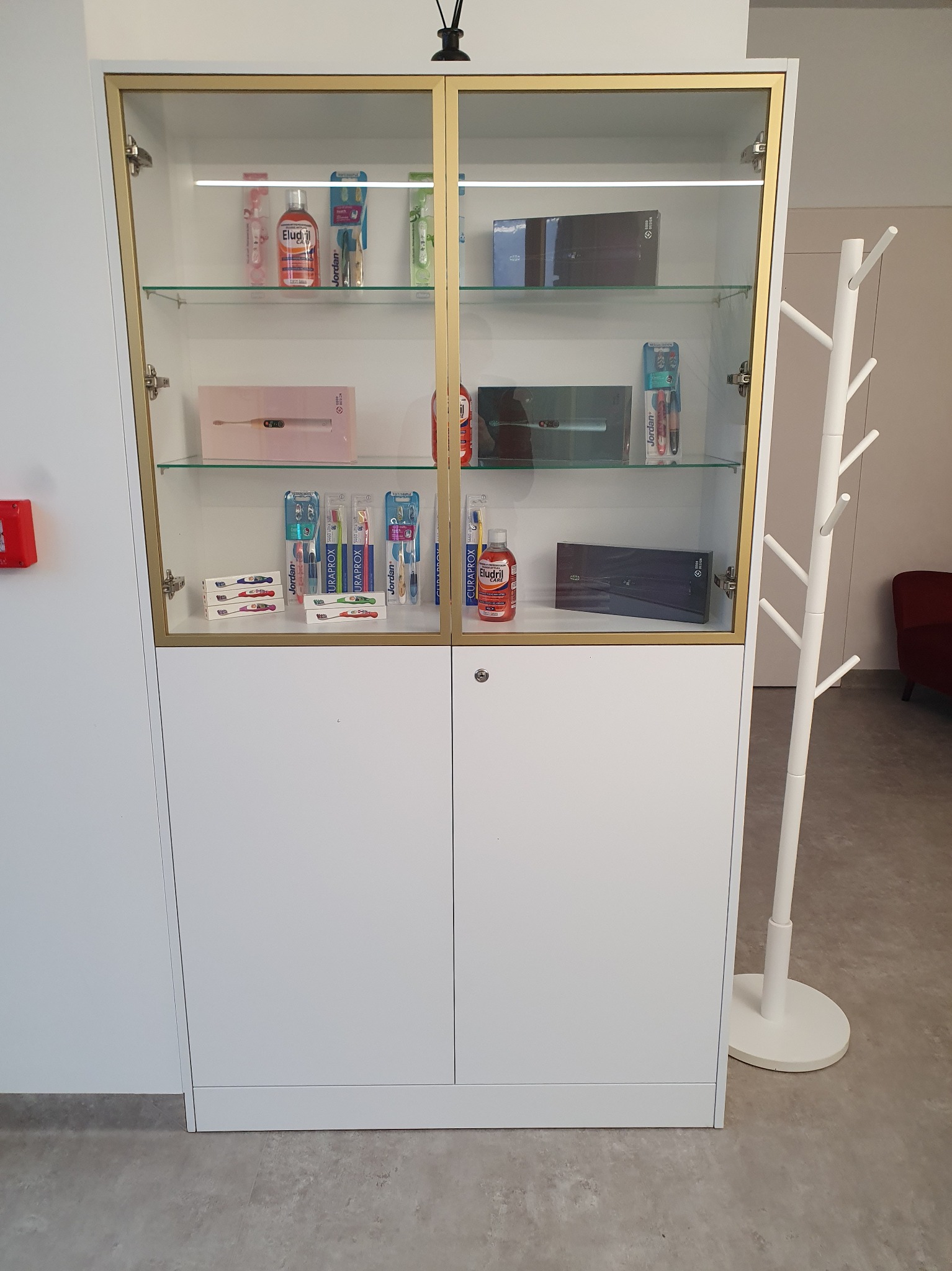 Biała szafka z przeszkloną, złotą ramą, prezentująca produkty do higieny jamy ustnej, obok wieszak na ubrania.