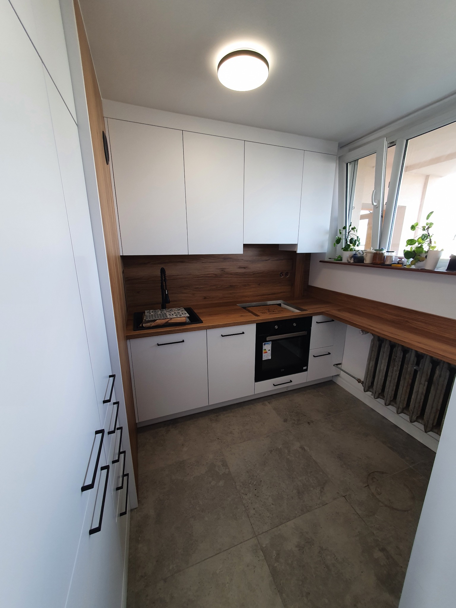 Nowoczesna, jasna kuchnia z białymi szafkami, drewnianym blatem i czarnymi uchwytami, zlew z czarną baterią, widok na parapet z roślinami w doniczkach, grzejnik żeliwny pod blatem.