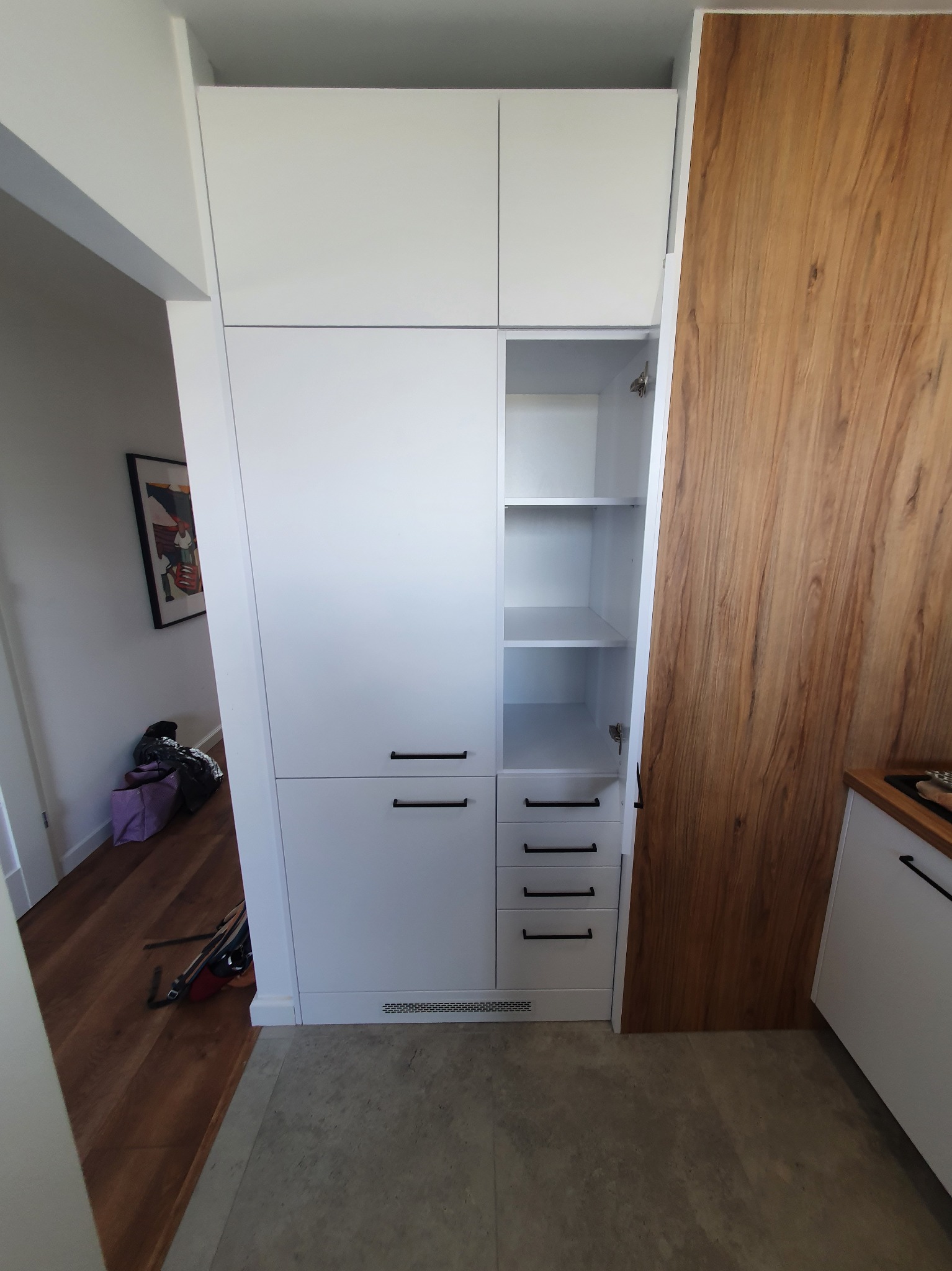 Wysoka biała szafa z czarnymi uchwytami, częściowo otwarta, z półkami, wkomponowana w przestrzeń obok drewnianej ściany i w pobliżu wejścia do pomieszczenia z obrazem na ścianie.