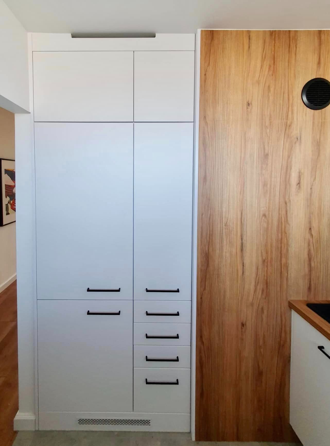 Biała zabudowa kuchenna z czarnymi, minimalistycznymi uchwytami, sąsiadująca z drewnopodobną okładziną ścienną z okrągłym otworem wentylacyjnym.