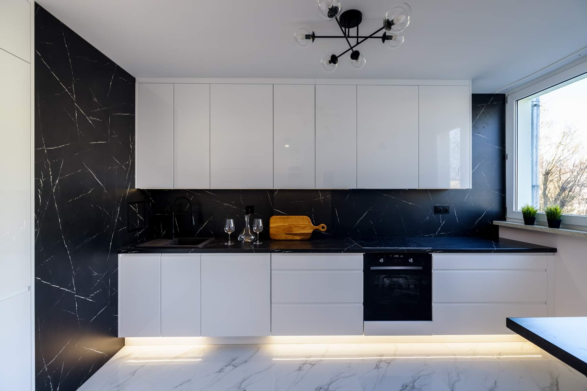 Nowoczesna kuchnia z białymi szafkami, czarnym blatem i marmurową ścianą, oświetlenie LED pod szafkami, drewniana deska do krojenia i szklane kieliszki na blacie.
