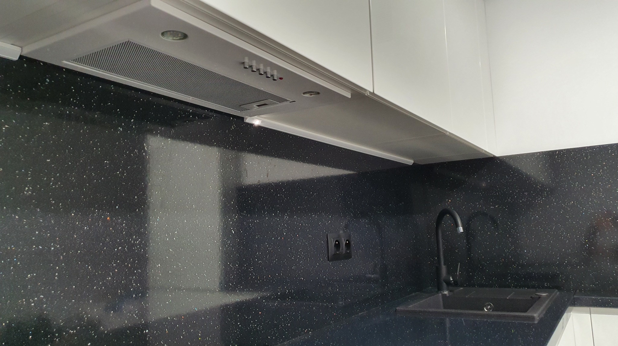 Fragment nowoczesnej kuchni z czarnym blatem imitującym gwiazdy, zlewozmywakiem z czarną baterią i białymi szafkami wiszącymi z okapem oraz oświetleniem podszafkowym.