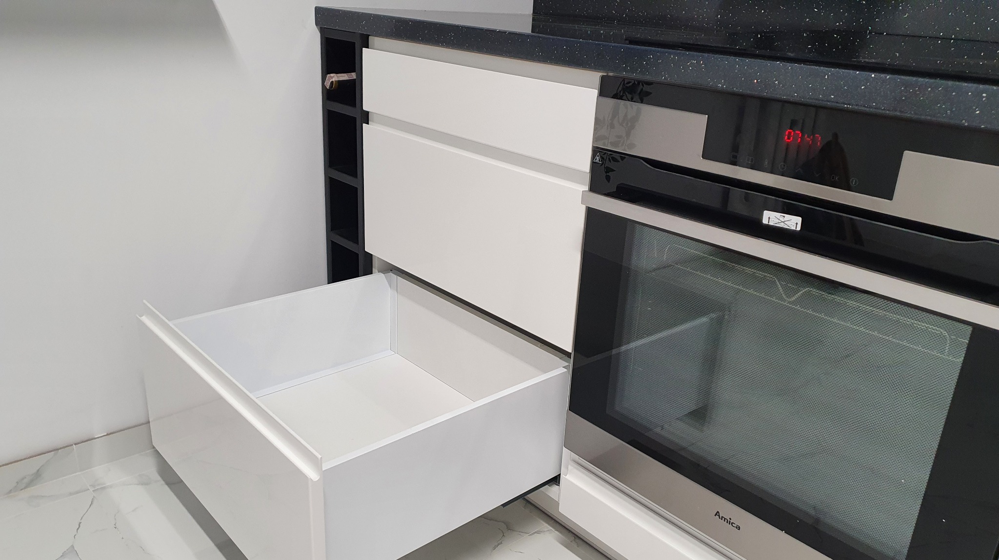Biała, minimalistyczna zabudowa kuchenna z otwartą szufladą i piekarnikiem Amica, czarny blat z brokatem, podłoga z marmurowym wzorem.