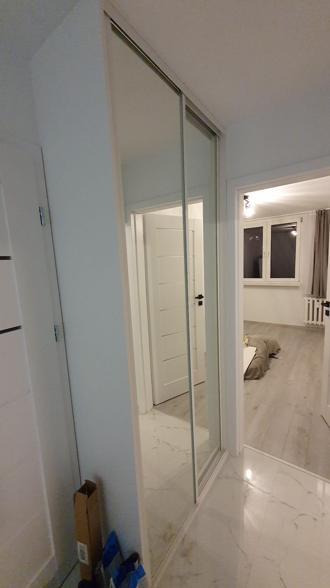 Nowoczesna szafa wnękowa z przesuwnymi drzwiami lustrzanymi, odbijająca wnętrze jasnego mieszkania z białymi drzwiami i marmurową podłogą.