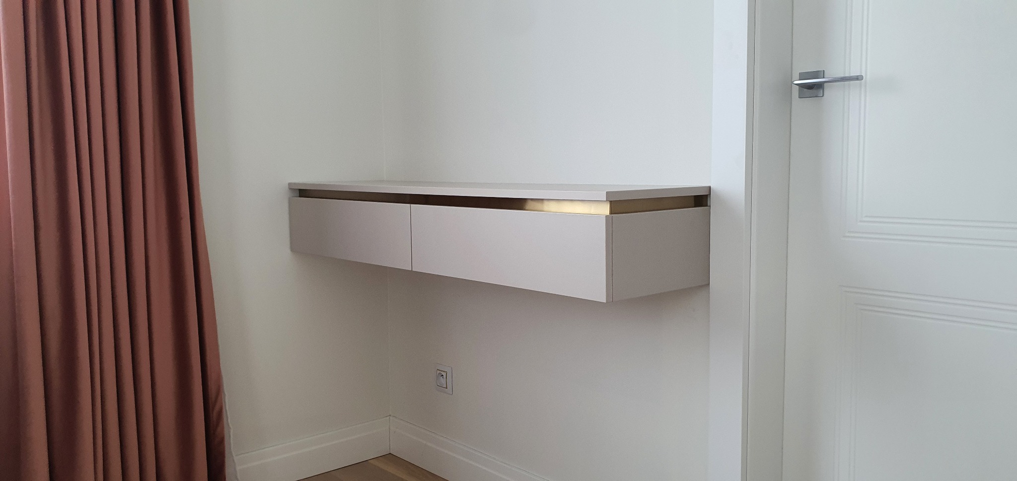 Biała, wisząca szafka z dwiema szufladami i złotym akcentem, zamontowana na ścianie obok białych drzwi i brązowej zasłony.