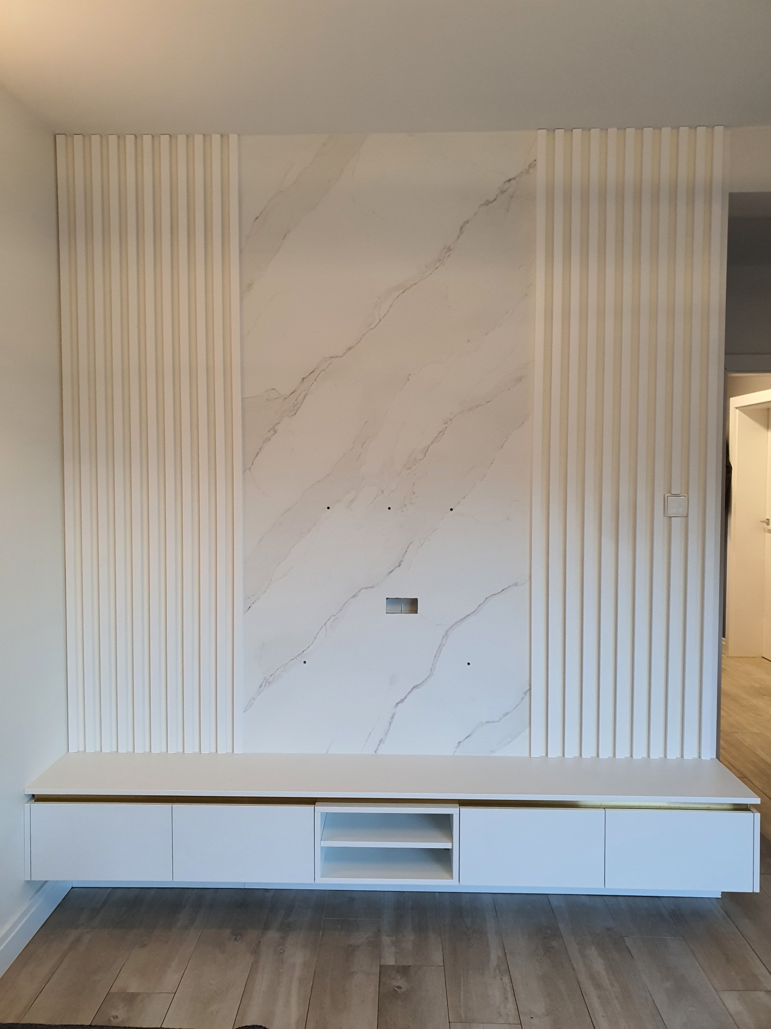 Biała zabudowa ściany telewizyjnej z marmurowym panelem i ryflowanymi listwami, podwieszana szafka RTV z subtelnym złotym akcentem.