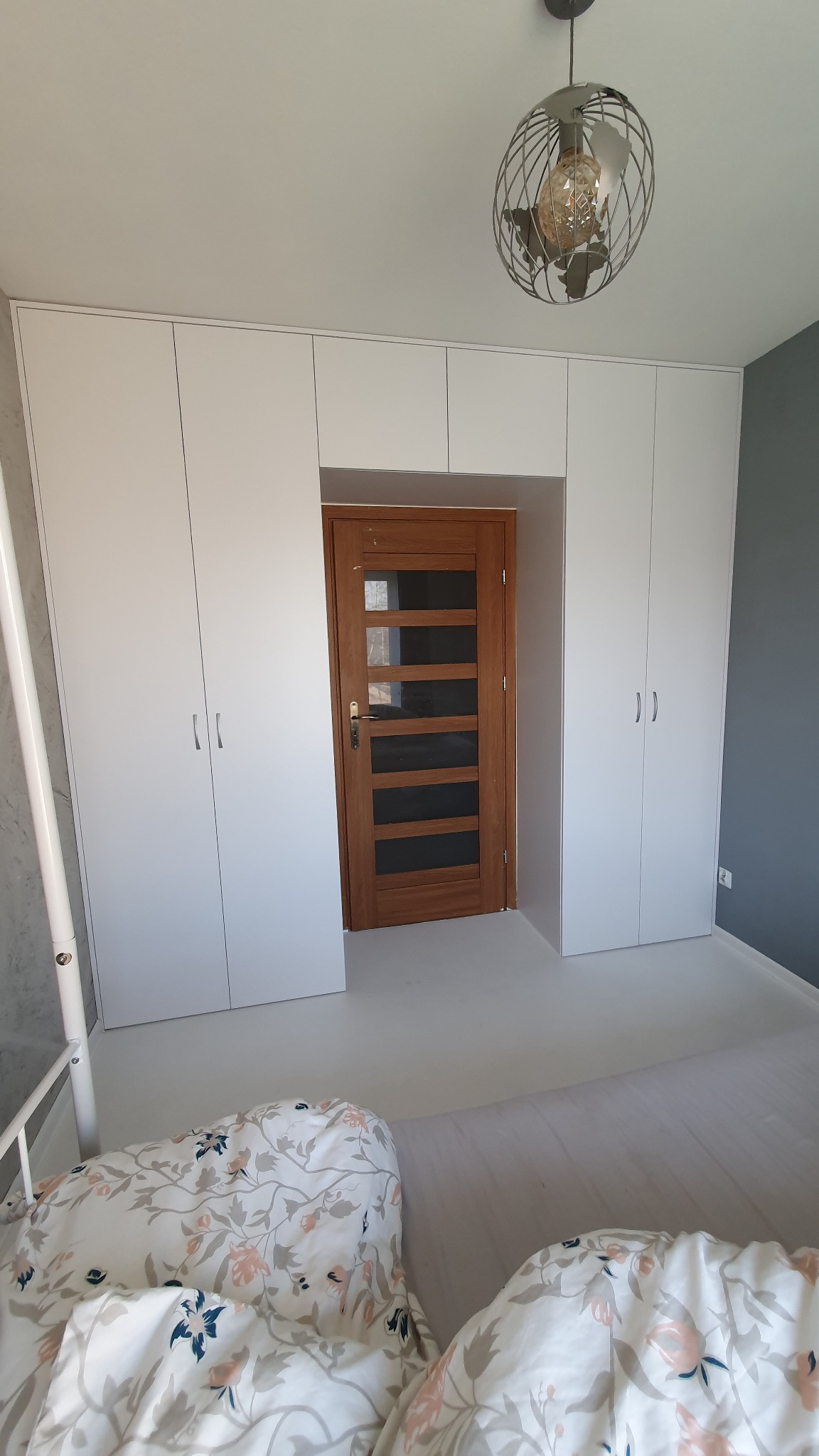 Białe szafy wnękowe z drewnianą, przeszkloną wstawkami, drzwiami, wkomponowane we wnękę, widok z perspektywy łóżka z pościelą w kwiaty, w tle szara ściana i lampa w stylu industrialnym.