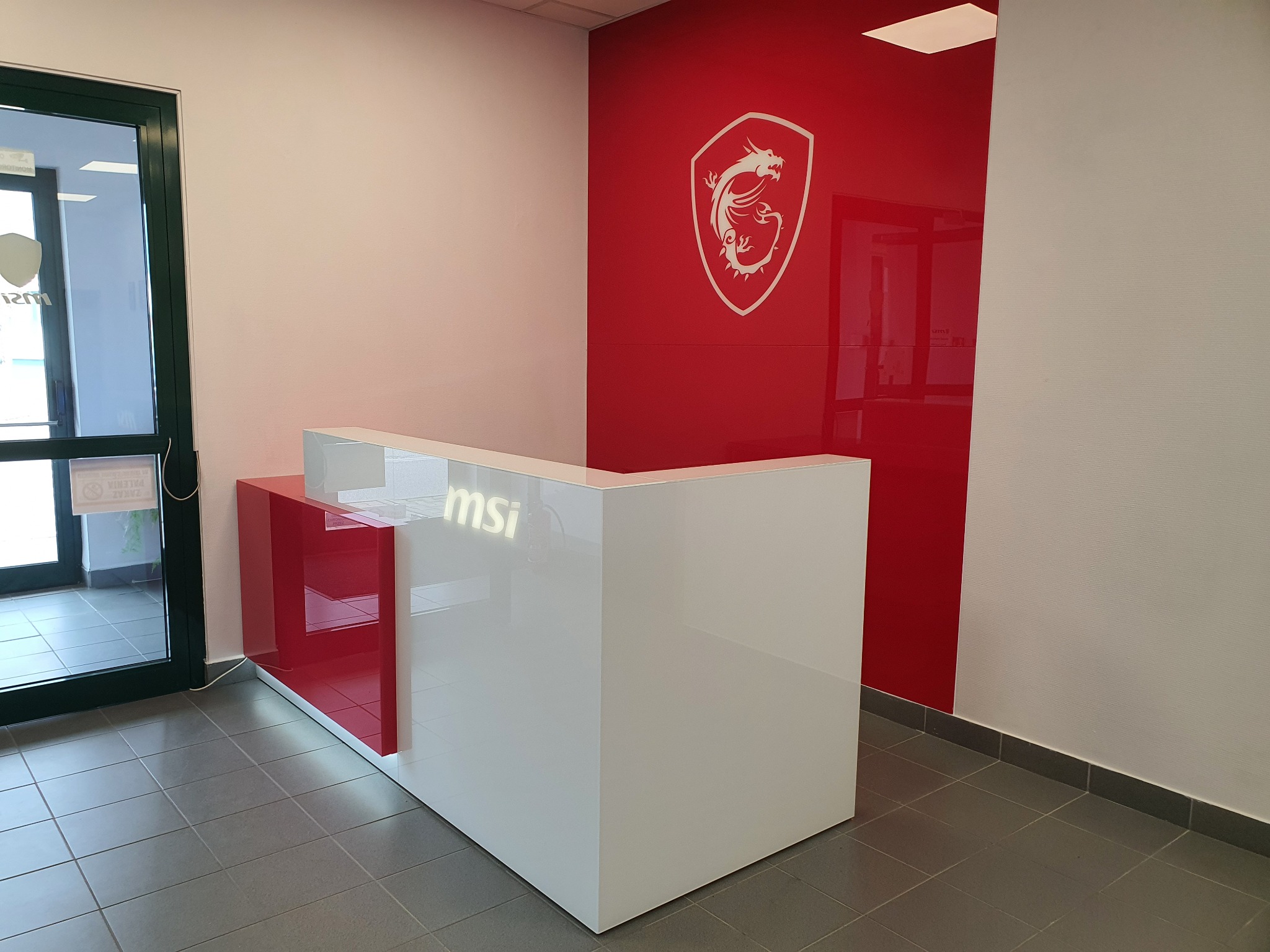 Nowoczesna recepcja z białym biurkiem i czerwonym akcentem, logo firmy MSI na biurku i ścianie, szara podłoga z płytek.