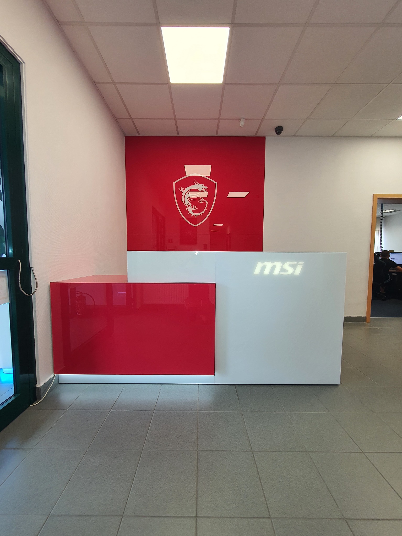 Nowoczesna recepcja w biurze z białym blatem i czerwonym panelem bocznym, logo firmy MSI na ścianie i blacie, widok na salę z pracownikami w tle.