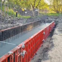 Wylany beton w szalunku zbrojonym prętami stalowymi, widok z boku na placu budowy w słoneczny dzień.