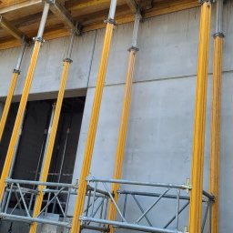MG Inżynieria Marek Grabias - Szare betonowe ściany budynku w trakcie budowy, podparte żółtymi stemplami budowlanymi i metalowym rusztowaniem, widoczne otwory okienne.