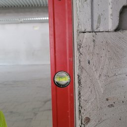 MG Inżynieria Marek Grabias - Czerwona poziomica przyłożona do surowej ściany z betonu komórkowego podczas prac budowlanych wewnątrz budynku.