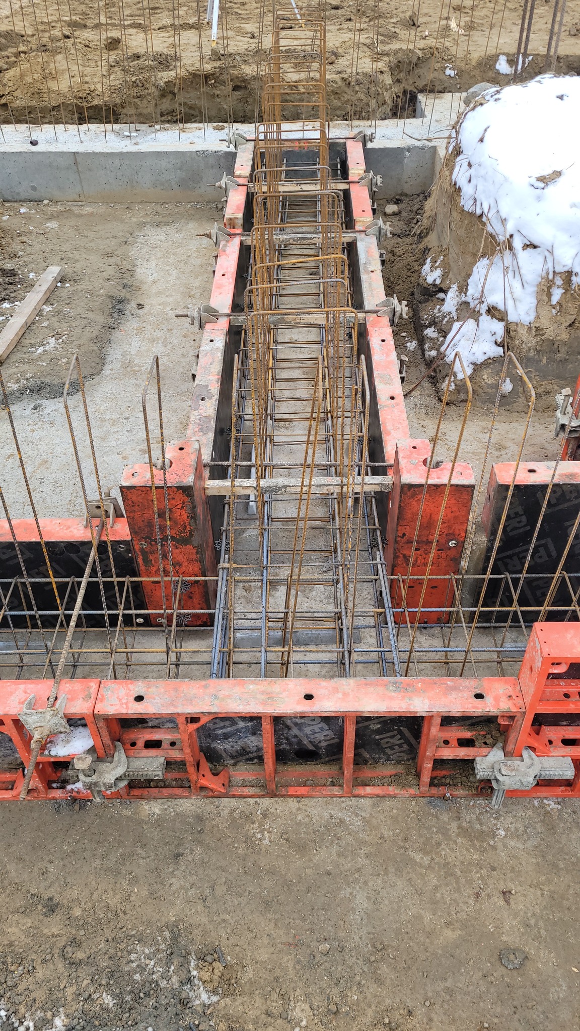 Zbrojenie fundamentu w szalunkach systemowych, widoczne stalowe pręty i elementy mocujące, ziemia z resztkami śniegu w tle.