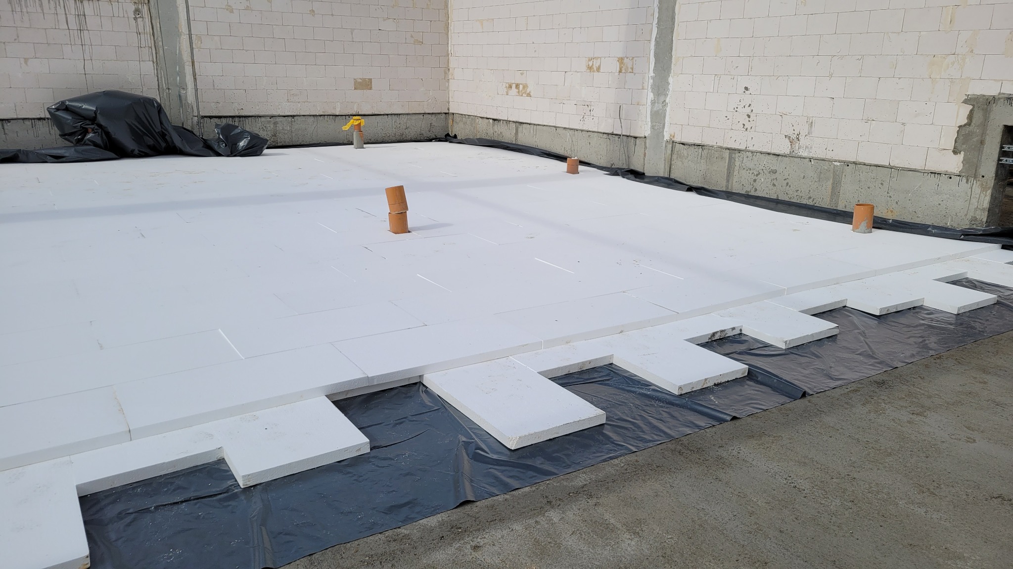 Układanie białych płyt izolacyjnych na czarnej folii w surowym wnętrzu budynku, widoczne rury instalacyjne wystające z podłoża.