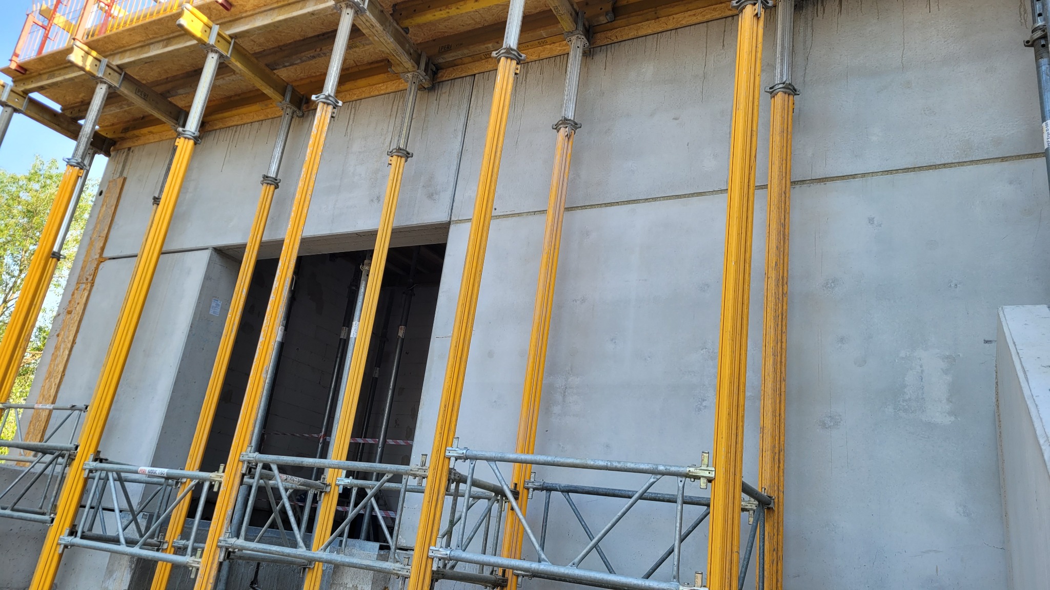 Szare betonowe ściany budynku w trakcie budowy, podparte żółtymi stemplami budowlanymi i metalowym rusztowaniem, widoczne otwory okienne.