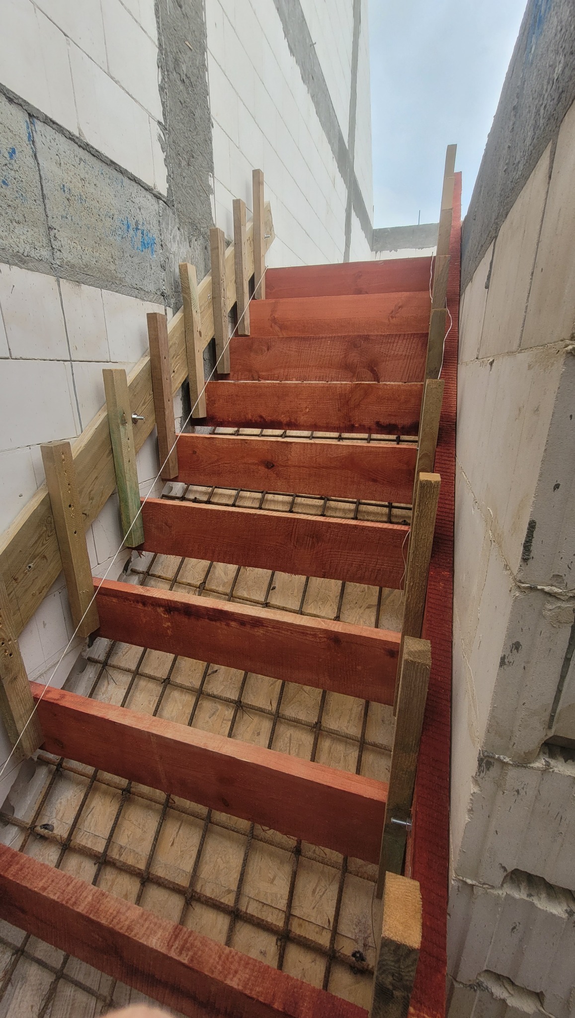Konstrukcja schodów żelbetowych w trakcie budowy: drewniane szalunki i zbrojenie stalowe na tle ściany z bloczków betonowych.