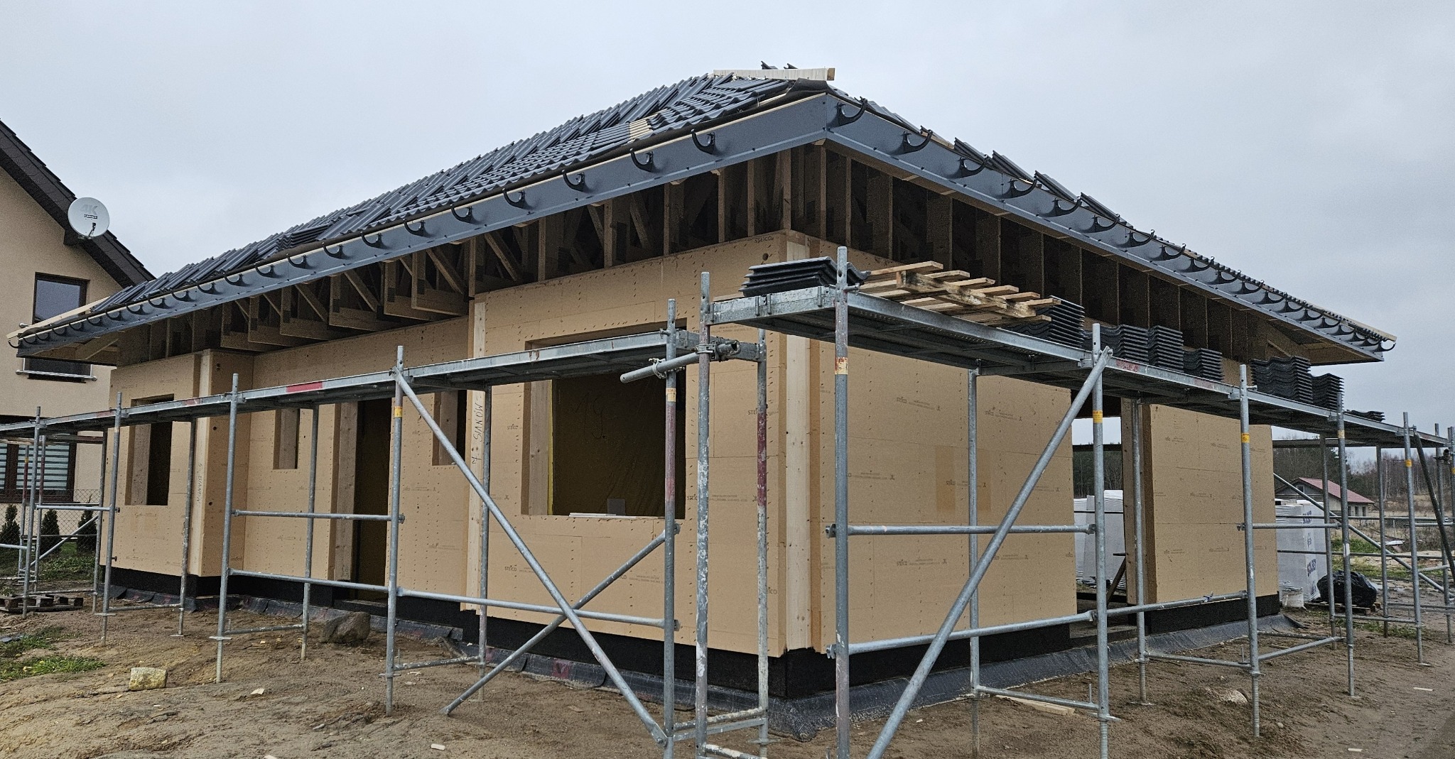 Dom w trakcie budowy z drewnianych paneli, widoczne rusztowania i dach z czarną dachówką. Ujęcie z zewnątrz, w pochmurny dzień.