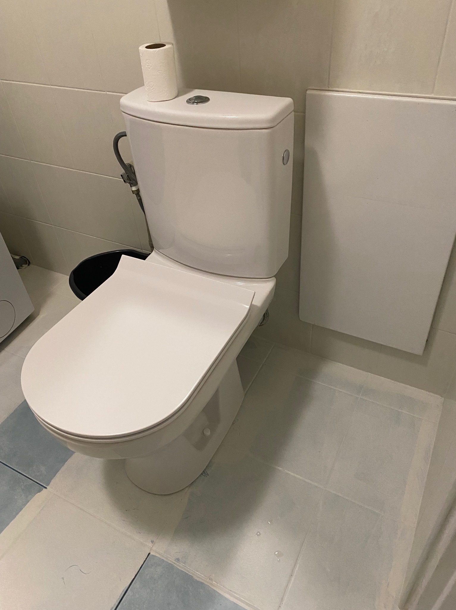 Biała toaleta kompaktowa z rolką papieru toaletowego na zbiorniku, widoczne przyłącze wody i fragment niebiesko-białej podłogi w łazience.