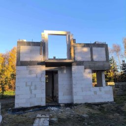 Usługi ogólno budowlane Jakub Łupicki - Surowy stan dwukondygnacyjnego domu z betonu komórkowego, widoczny otwór na okno i drzwi, niebieski wąż na ziemi, jesienne drzewa w tle.