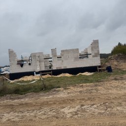 Usługi ogólno budowlane Jakub Łupicki - Wznoszone ściany parteru domu z białych bloczków na fundamencie zabezpieczonym czarną izolacją, widoczne rusztowania i teren budowy w pochmurny dzień.