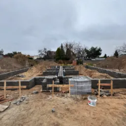 Budowa fundamentów domu z bloczków betonowych, widoczne zbrojenia, szalunki drewniane i stosy bloczków na ziemi, ziemia wokół wykopu.