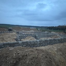 Usługi ogólno budowlane Jakub Łupicki - Wykop i fundamenty pod budowę domu jednorodzinnego z bloczków betonowych, widoczny stos bloczków, ziemia i zarys lasu w tle przy pochmurnej pogodzie.