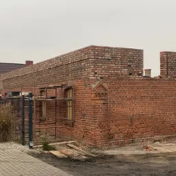 Wznoszenie muru z czerwonej cegły z widocznymi rusztowaniami i fragmentem dachu sąsiedniego budynku w tle.