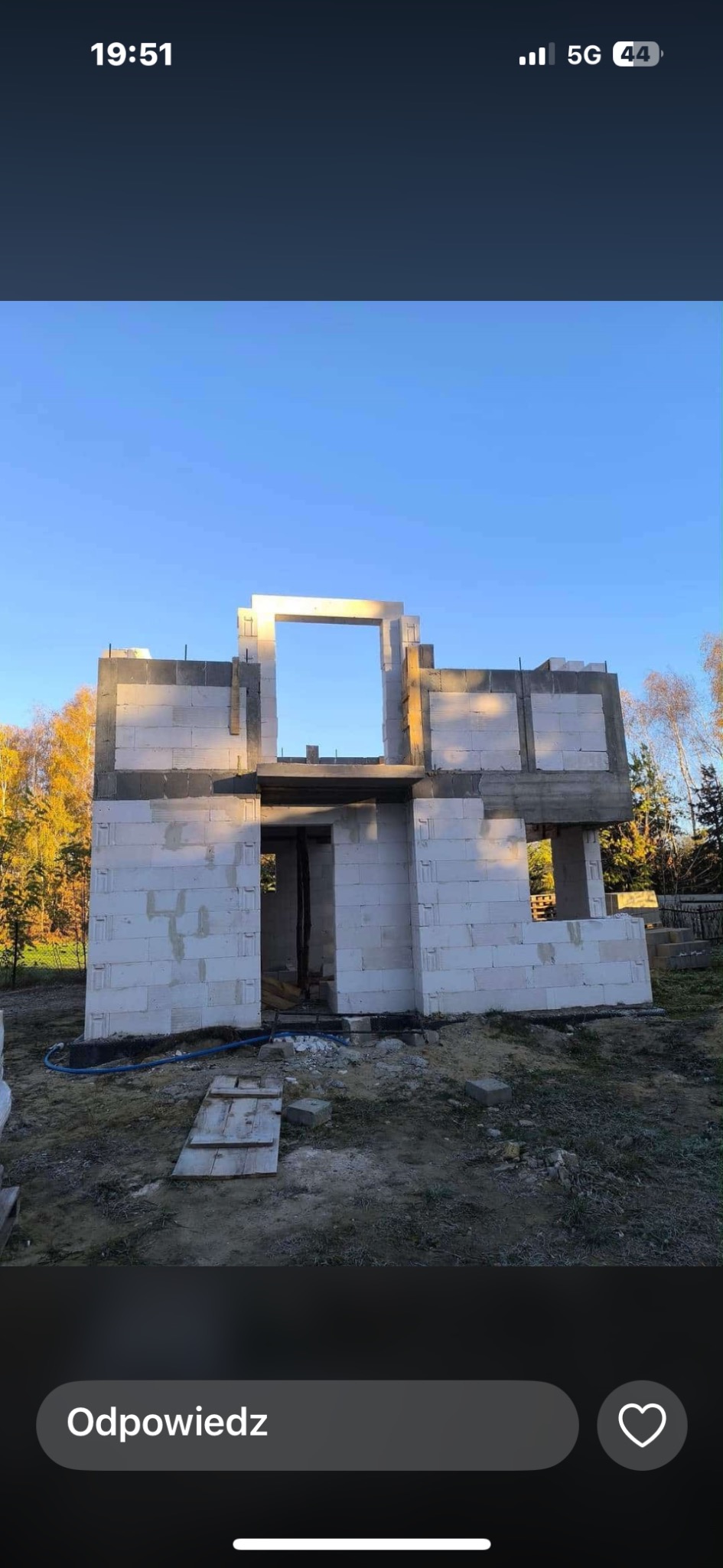 Surowy stan dwukondygnacyjnego domu z betonu komórkowego, widoczny otwór na okno i drzwi, niebieski wąż na ziemi, jesienne drzewa w tle.