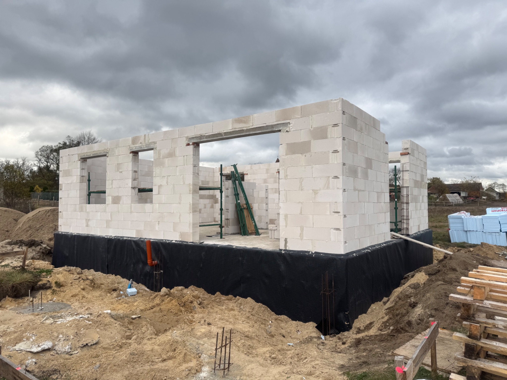 Wznoszenie ścian parterowego domu z białych bloczków, widoczne zbrojenia i izolacja fundamentów, drabina oparta o ścianę, pochmurne niebo.