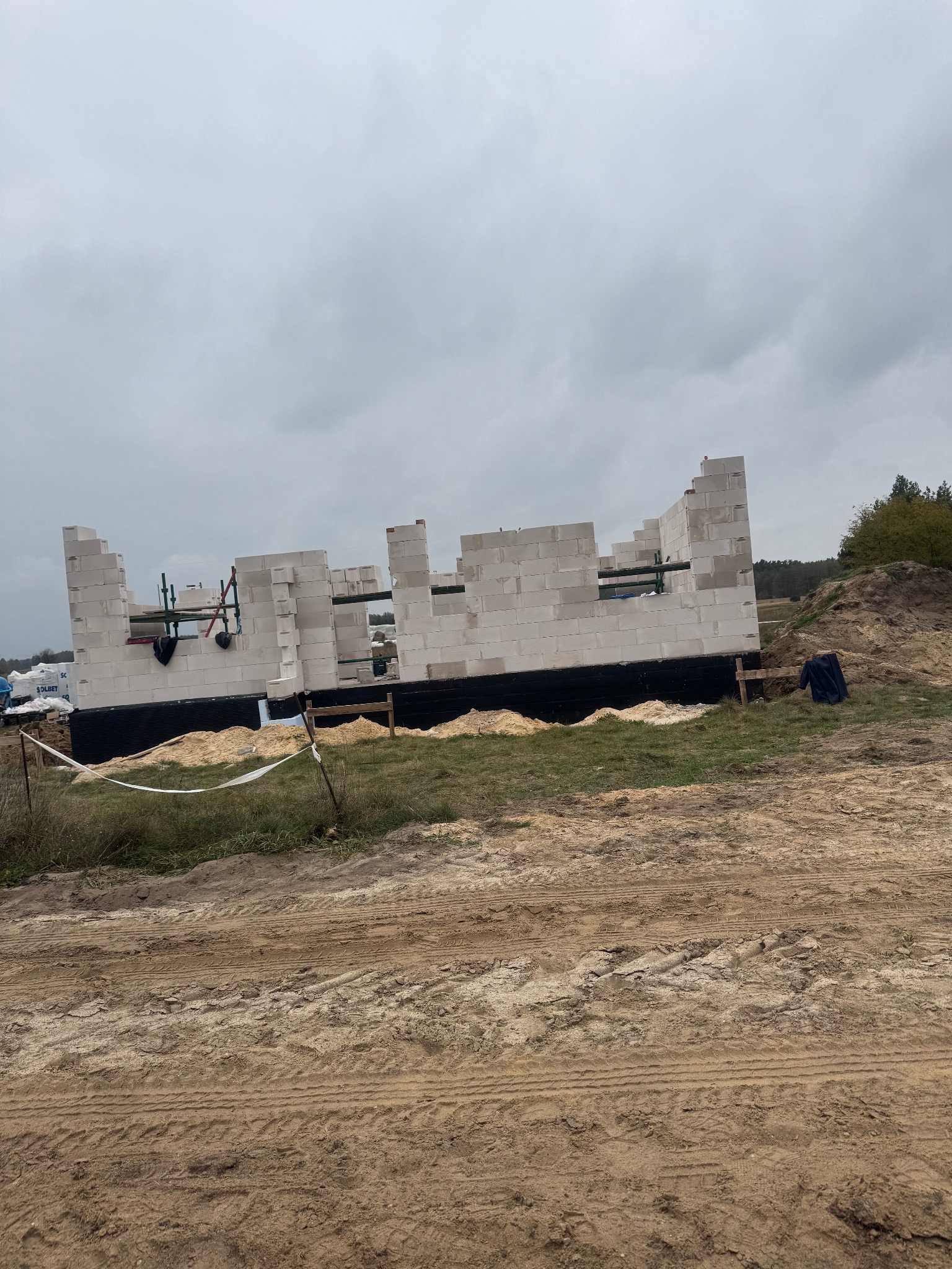 Wznoszone ściany parteru domu z białych bloczków na fundamencie zabezpieczonym czarną izolacją, widoczne rusztowania i teren budowy w pochmurny dzień.
