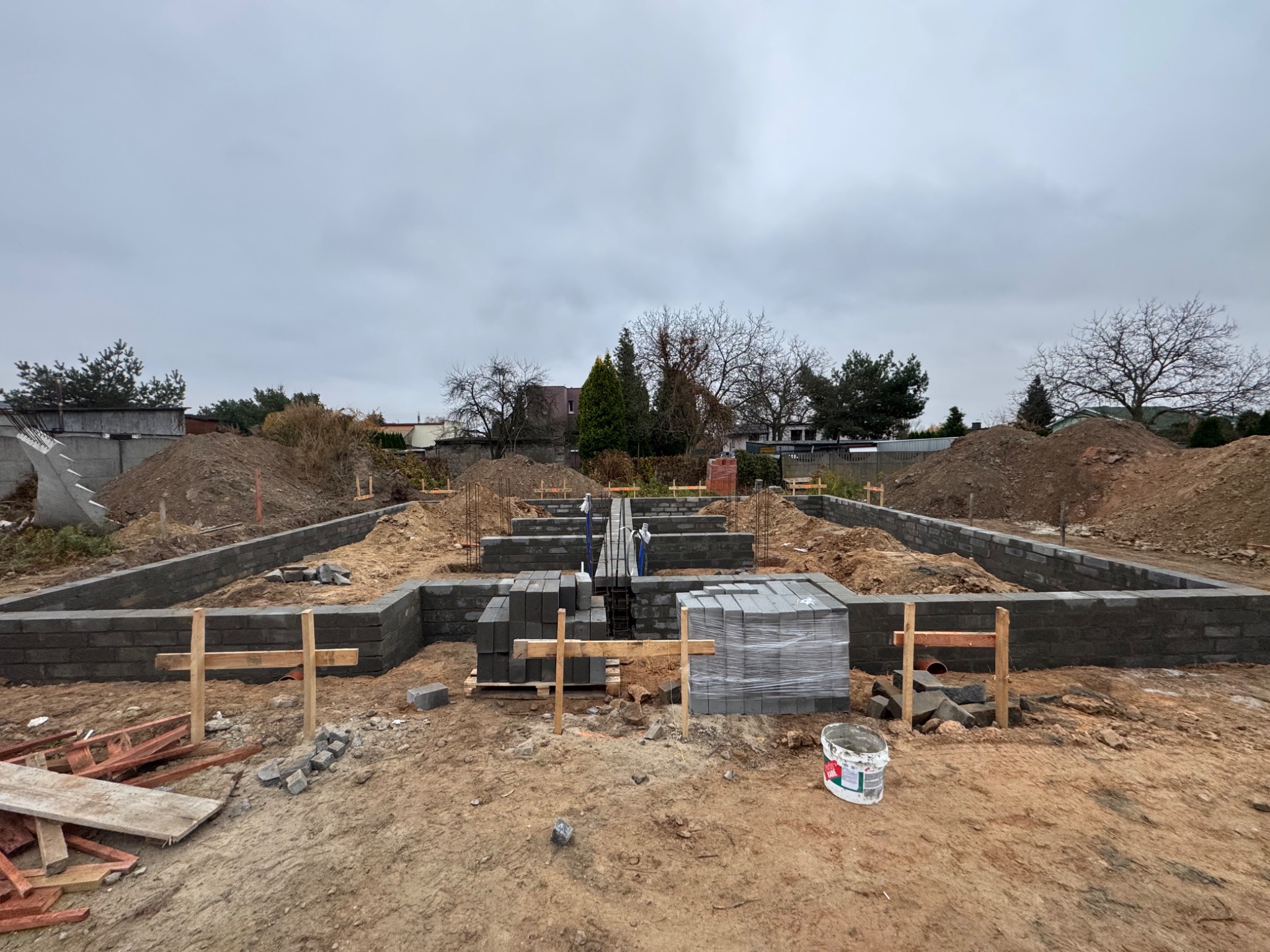 Budowa fundamentów domu z bloczków betonowych, widoczne zbrojenia, szalunki drewniane i stosy bloczków na ziemi, ziemia wokół wykopu.