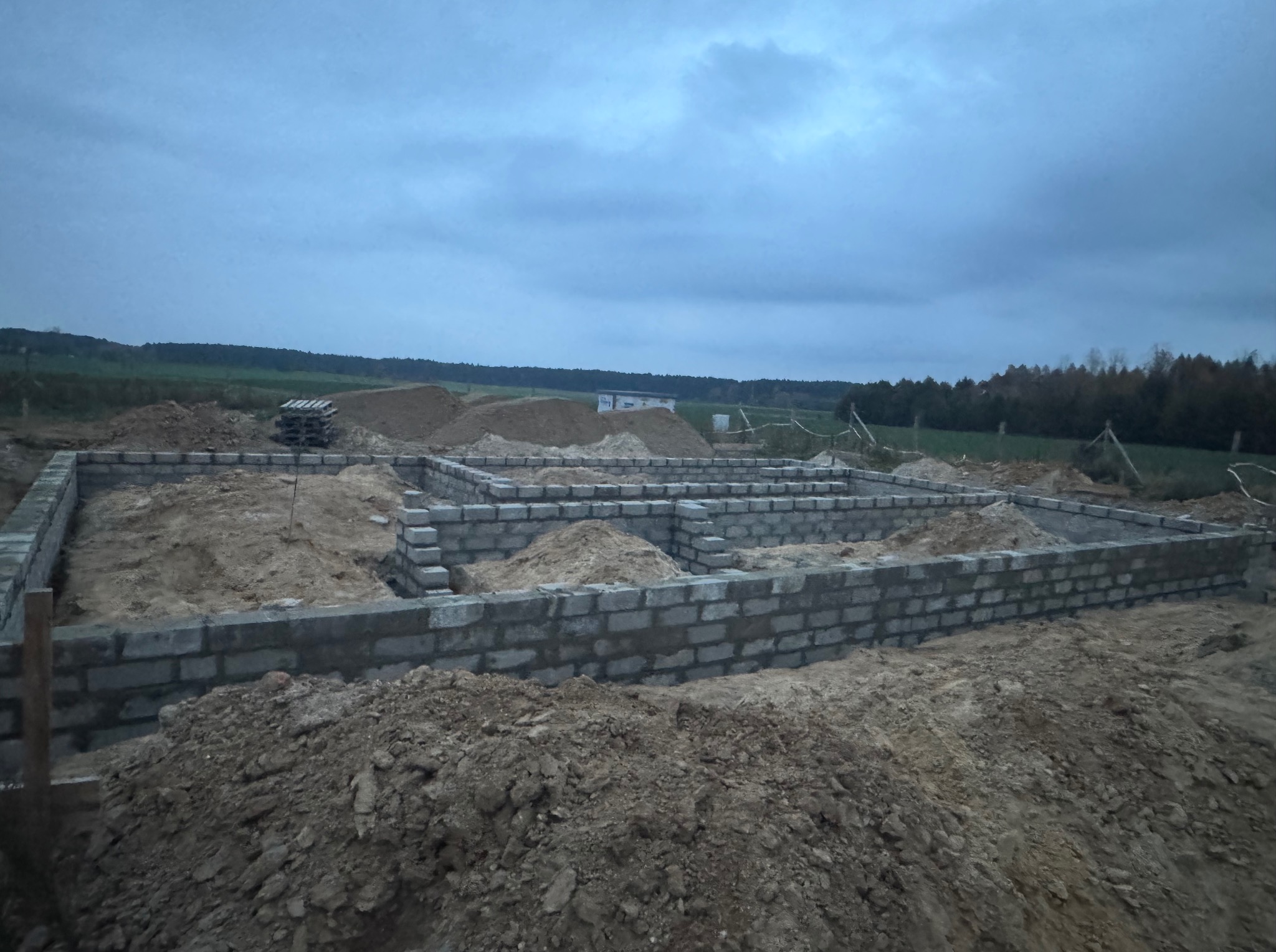 Wykop i fundamenty pod budowę domu jednorodzinnego z bloczków betonowych, widoczny stos bloczków, ziemia i zarys lasu w tle przy pochmurnej pogodzie.