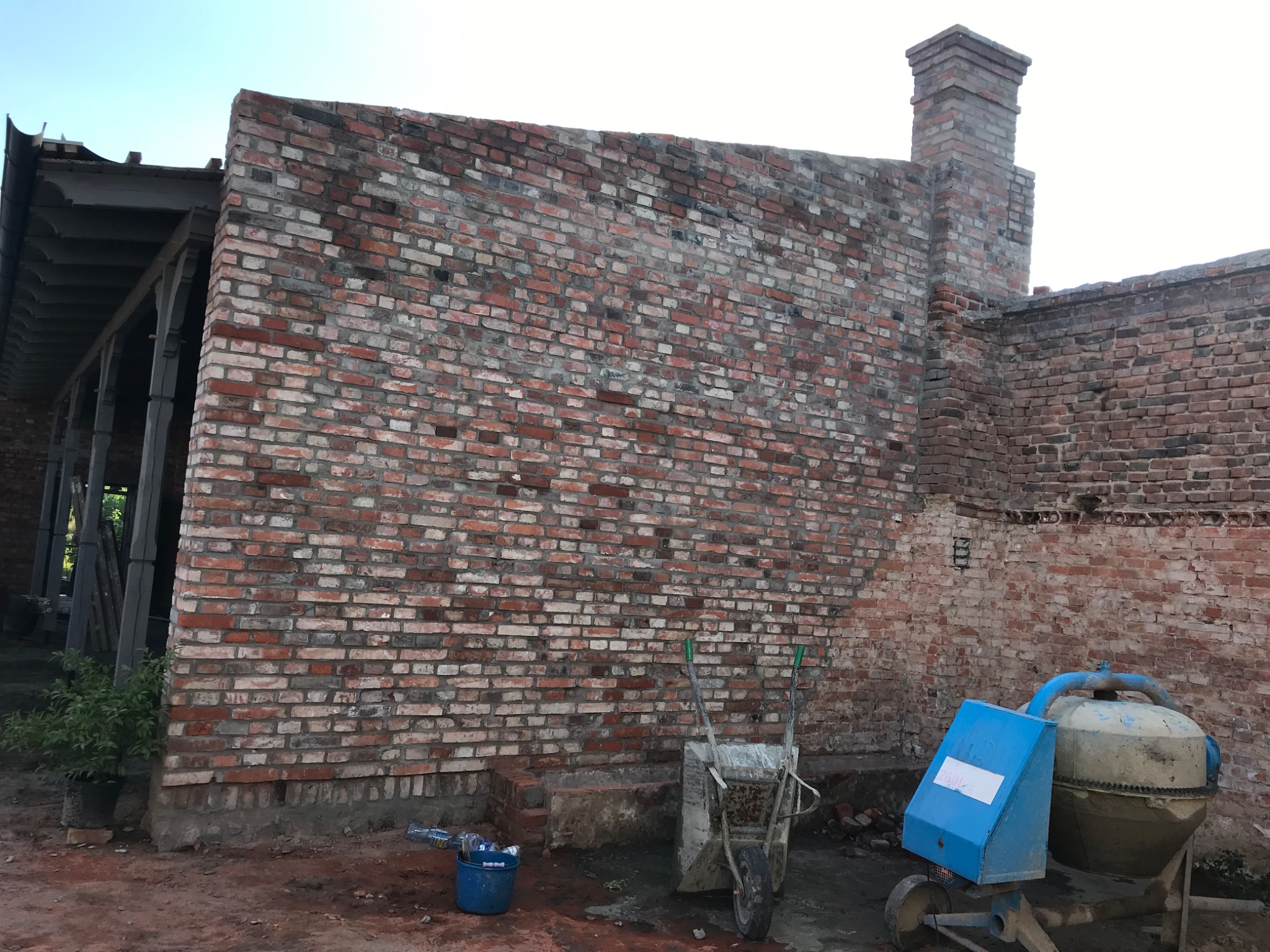 Wznoszenie ściany z czerwonej cegły z widoczną zaprawą, obok stoi taczka i brudna betoniarka, w tle zadaszona konstrukcja.