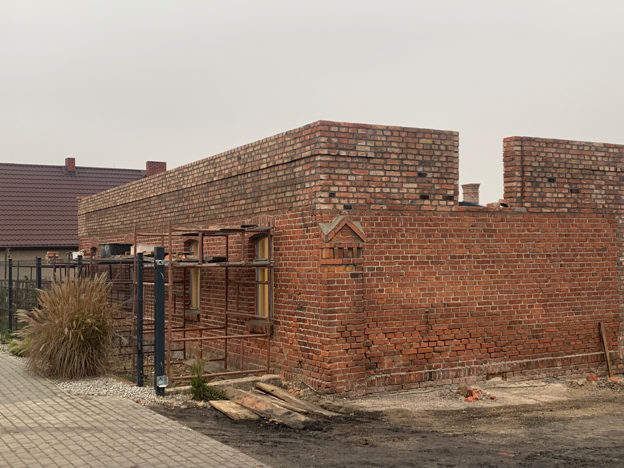 Wznoszenie muru z czerwonej cegły z widocznymi rusztowaniami i fragmentem dachu sąsiedniego budynku w tle.
