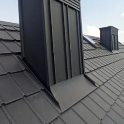 Czarny, metalowy komin z poziomymi żaluzjami na szczycie, wystający z ciemnego dachu krytego blachodachówką na tle błękitnego nieba.