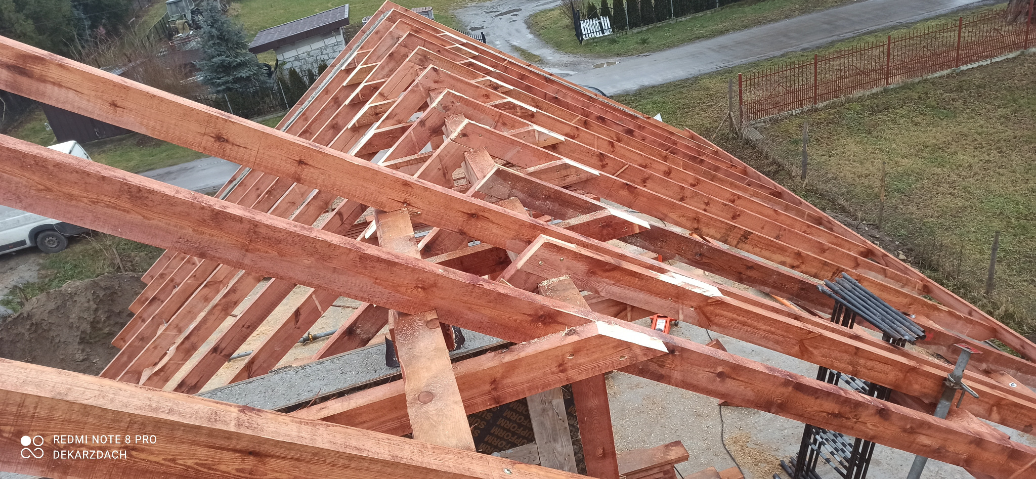 Konstrukcja drewniana dachu w trakcie budowy, widok z góry, elementy impregnowane na brązowo, widoczne narzędzia i elementy montażowe, tło stanowi krajobraz wiejski w pochmurny dzień.