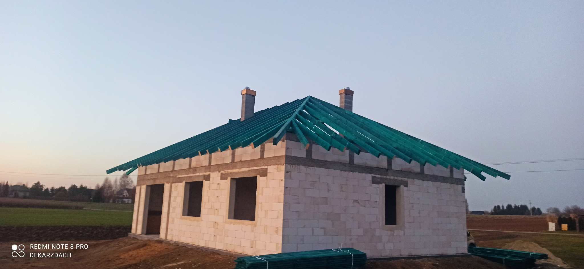 Niedokończony dom z widoczną konstrukcją drewnianą dachu, przygotowany do dalszych prac dekarskich, w tle krajobraz wiejski o zmierzchu.