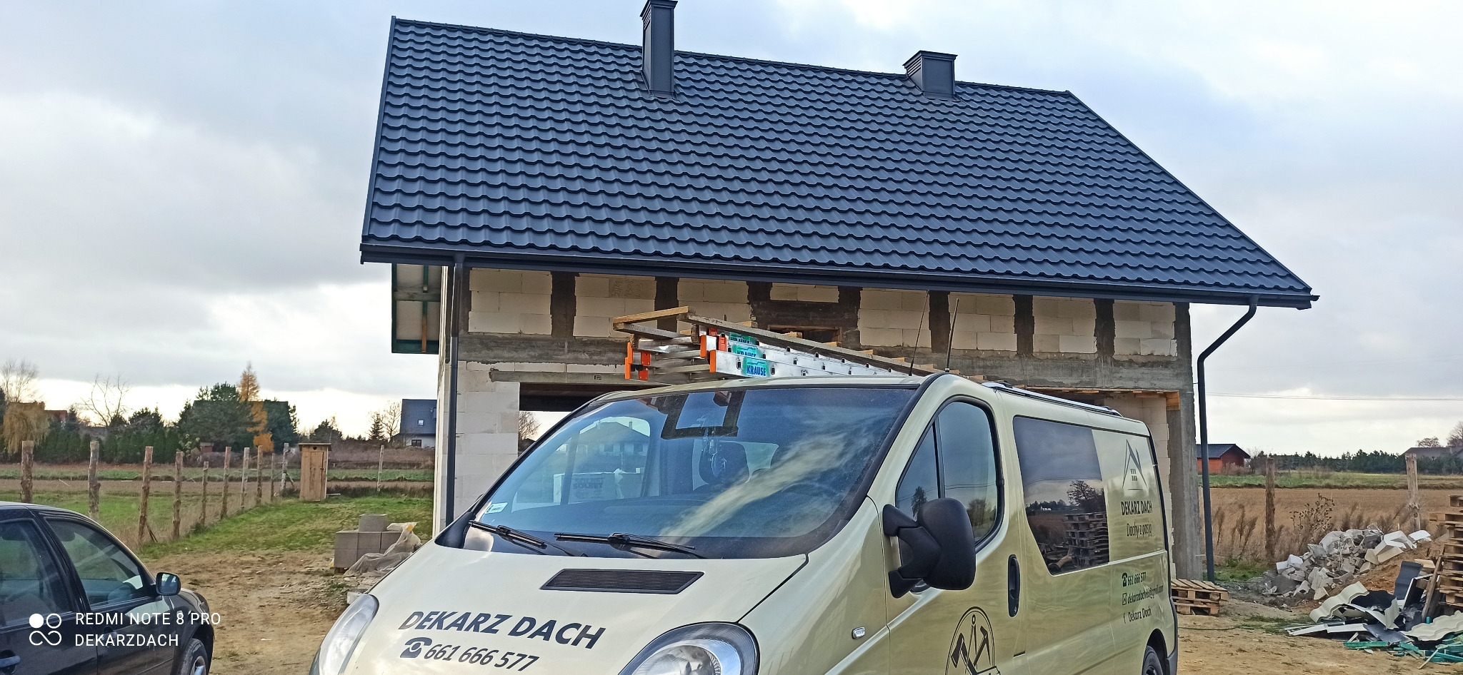 Nowy, ciemny dach z blachy na surowym budynku z pustaków, z widoczną rynną, drabina oparta o ścianę, firmowy samochód dekarski na pierwszym planie, jesienny krajobraz w tle.