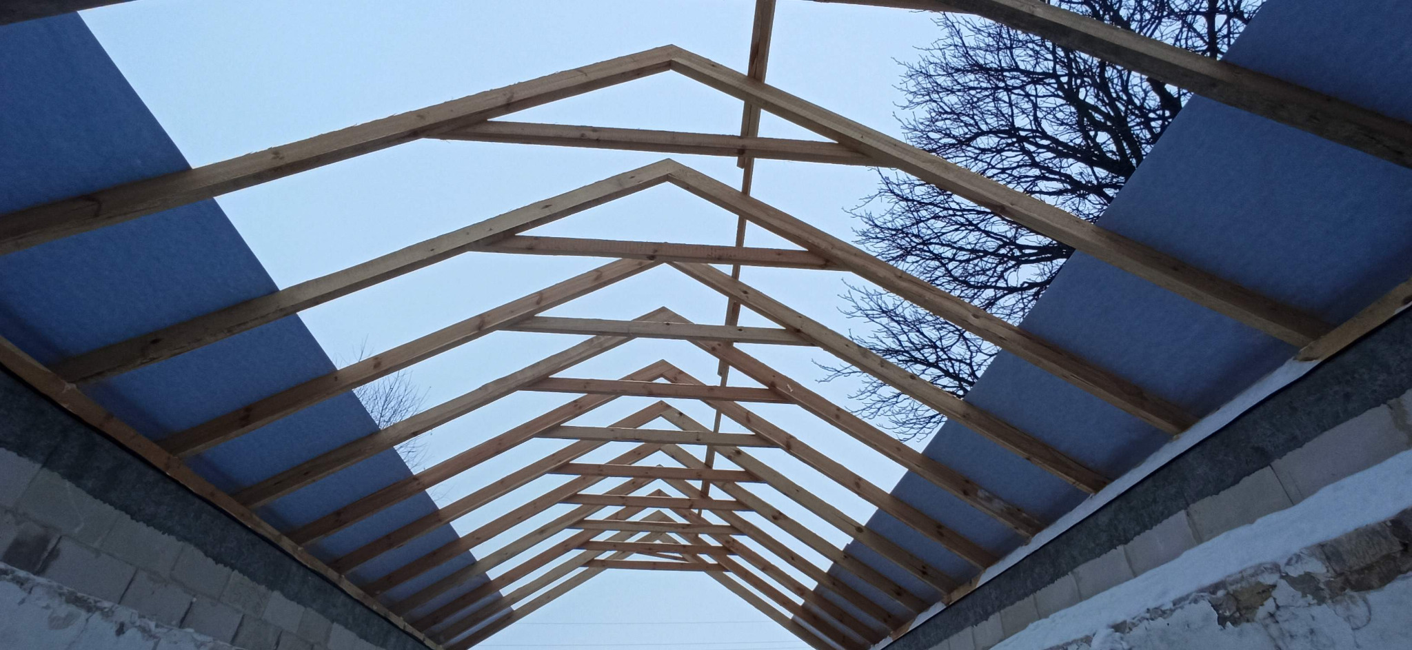 Konstrukcja drewniana dachu z widoczną membraną dachową, widok z wnętrza budynku w trakcie budowy, fragment muru z bloczków i drzewo bez liści na tle jasnego nieba.