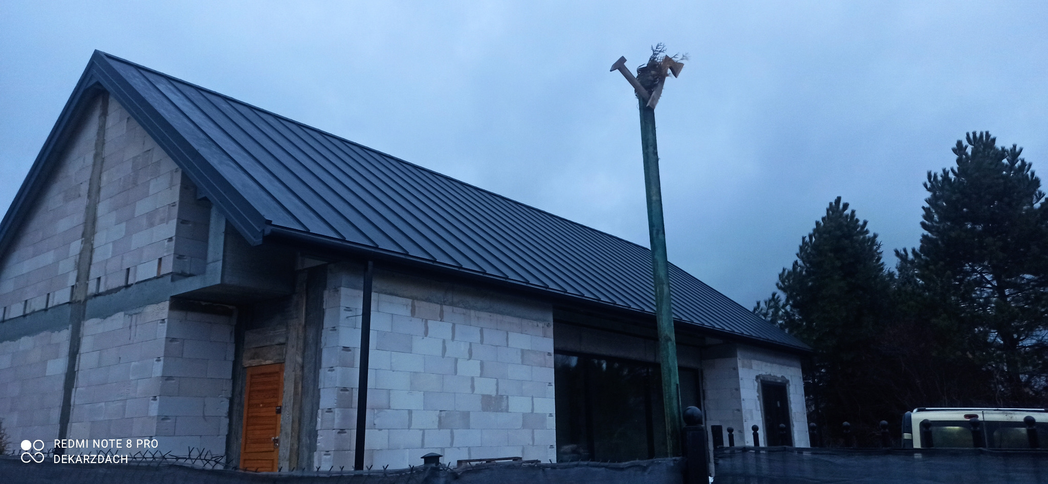 Nowoczesny dom w trakcie budowy z ciemnym metalowym pokryciem dachowym, widoczna surowa konstrukcja ścian z betonu komórkowego i nietypowa dekoracja na wysokim słupie z przodu budynku