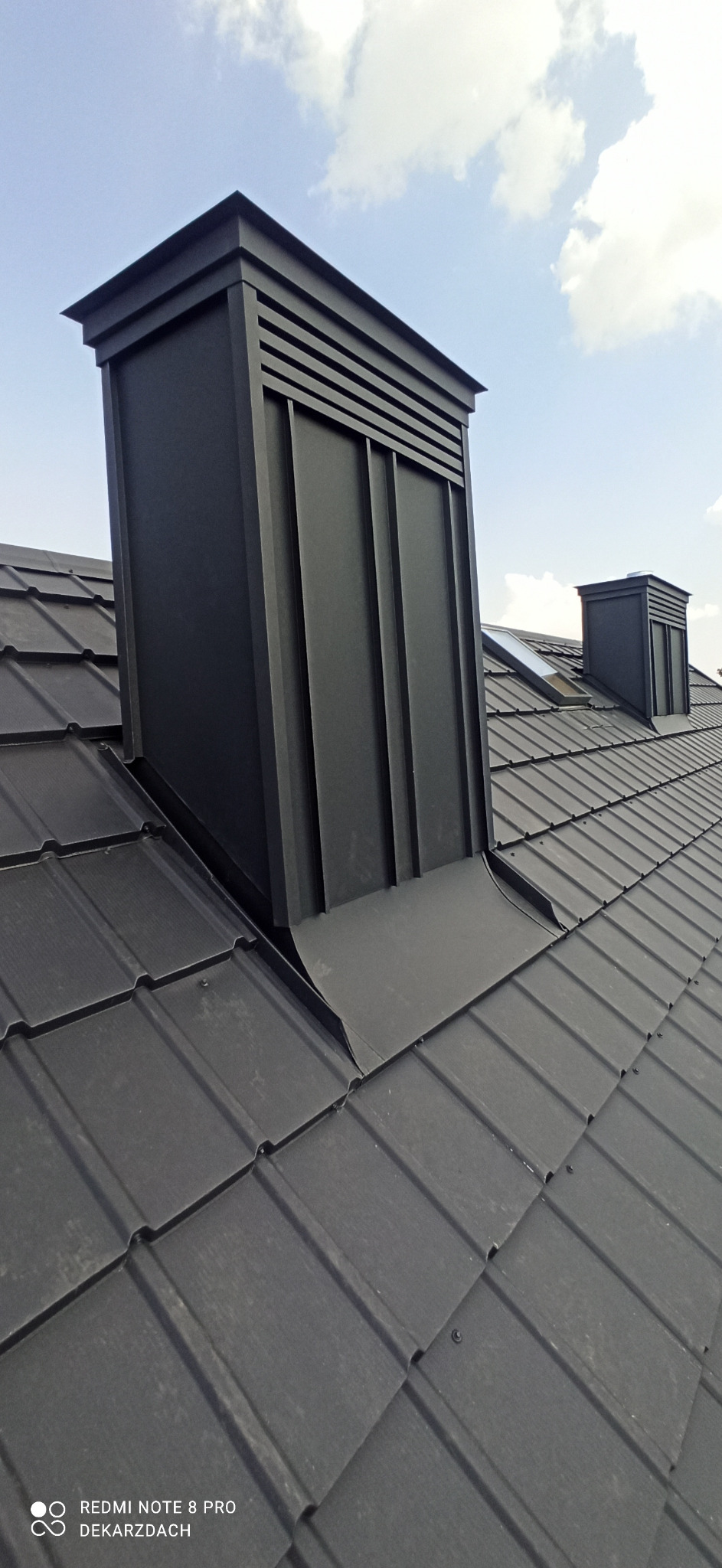 Czarny, metalowy komin z poziomymi żaluzjami na szczycie, wystający z ciemnego dachu krytego blachodachówką na tle błękitnego nieba.
